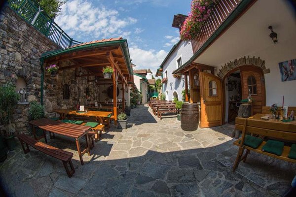 1_hoellmueller-innenhof-copyright-josef-salomon, © Josef Salomon Ein gemütlicher Innenhof mit Holzbänken, Tischen und Blumen. Steinboden und überdachte Sitzbereiche. Sonniges Wetter mit blauem Himmel.