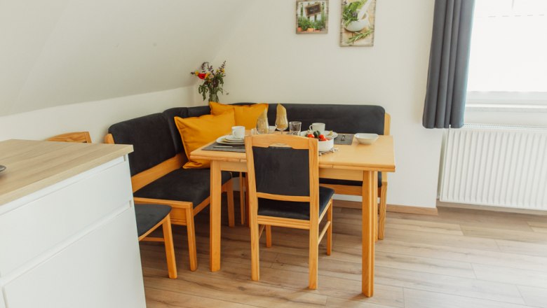 Appartement 2, © Ines Nejedly Essbereich mit Holztisch, Stühlen und Eckbank, gedeckt mit Geschirr, Blumenstrauß und Dekoration an der Wand.