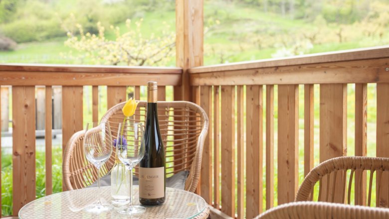 Ausblick vom Balkon, © Weingut Rixinger Ein Balkon mit Holzmöbeln, einer Weinflasche und Gläsern auf einem Tisch. Im Hintergrund grüne Landschaft.