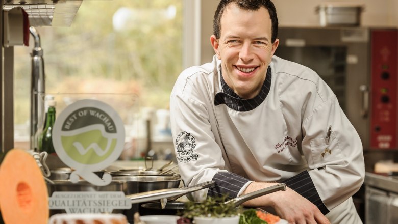 Master of the house, © Roman Siebenhandl A chef in a white uniform smiles in a kitchen surrounded by fresh ingredients and a Wachau quality seal.