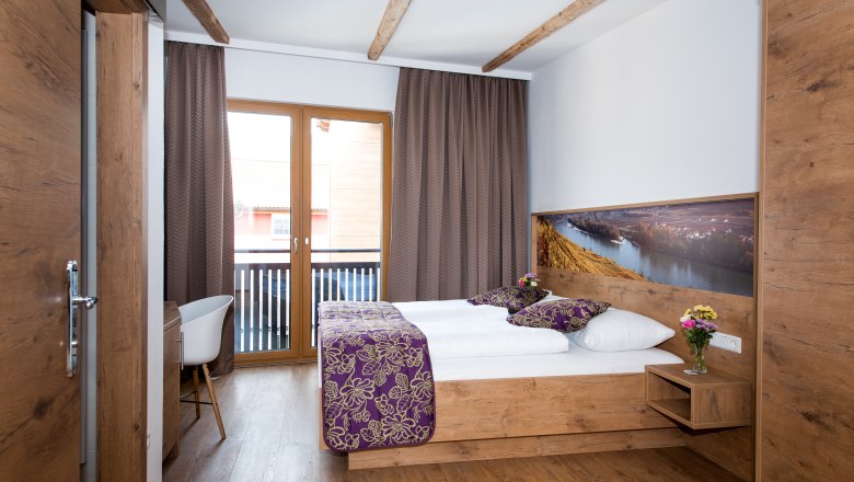 Room with balcony, © Martina Siebenhandl A cozy hotel room with wooden furniture, a double bed, balcony and decorative cushions.