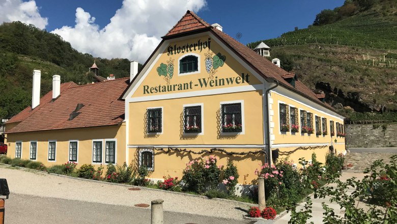 Exterior view of the monastery courtyard, © Restaurant Klosterhof Exterior view of a yellow building with the inscription 'Klosterhof Restaurant-Weinwelt', surrounded by flowers and vineyards.