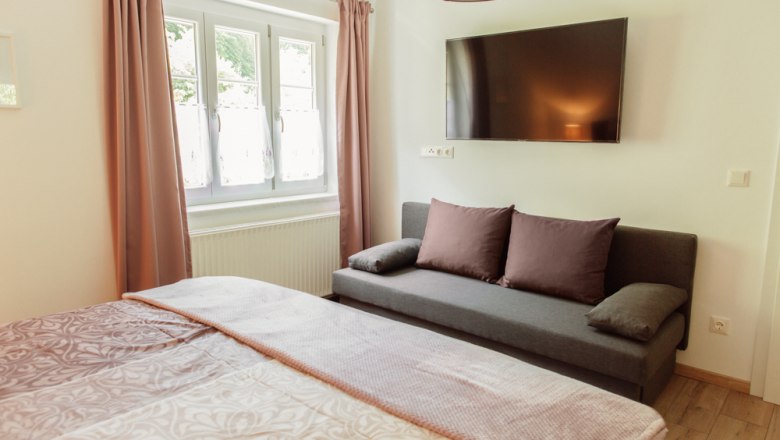 Appartement 2, © Ines Nejedly Innenansicht eines Schlafzimmers mit Bett, Sofa, Fenster und Wandfernseher.