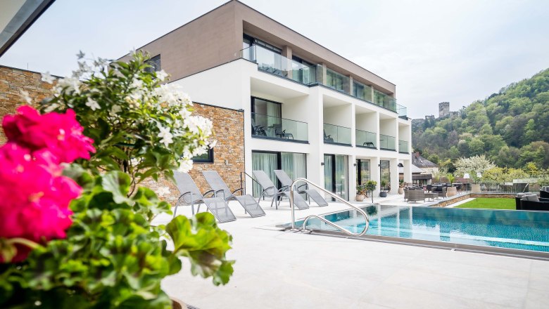 Donabaum © photography pfeffel, © © photography pfeffel Modernes Gebäude mit Pool und Liegestühlen, umgeben von grüner Landschaft und Burg im Hintergrund.