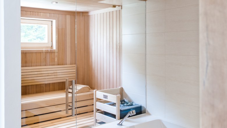 Sauna, © Martina Siebenhandl Interior view of a modern sauna with wooden paneling and glass wall, next to a white bathtub.
