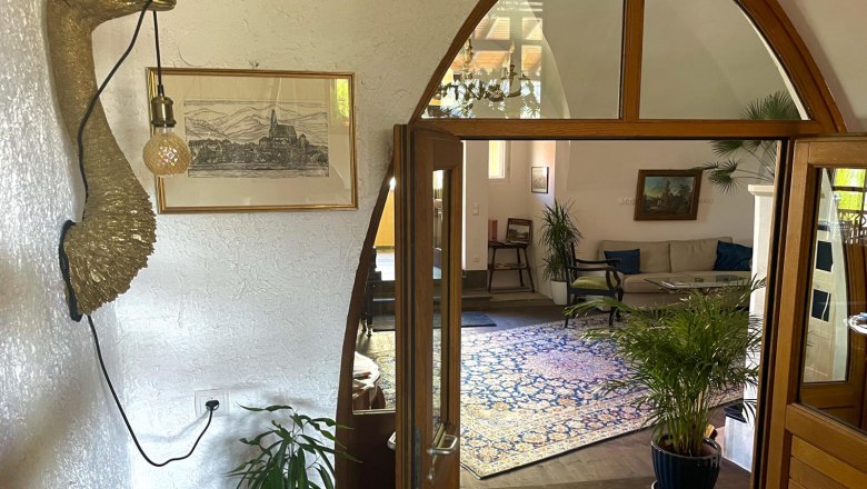 Departure, © Georg Smolka Interior view of a living room with plants, a sofa and a decorative wall object in the shape of a bird with a lamp.