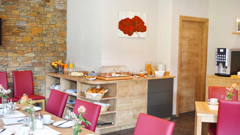 Breakfast buffet, © Familie Martin A breakfast buffet in a cozy room with red chairs, wooden furniture and a stone wall. The buffet includes croissants, muesli and orange juice.