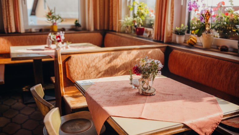 Gemütliche Stube für müde Wanderer, © Niederösterreich Werbung/Daniela Führer Gemütliche Stube mit Holzmöbeln, Tischdecken und Blumen auf den Tischen, umgeben von Fenstern mit Pflanzen.