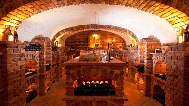 Wine cellar - Vinotheque, © Familie Pichler An atmospherically lit wine cellar with brick walls and wine bottles on shelves.