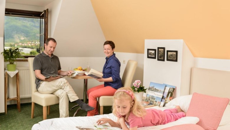 Zimmer, © Hamberger Familie in einem gemütlichen Zimmer, Eltern lesen am Tisch, Kind liegt auf dem Bett mit einem Buch.