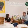 Gästezimmer, © RN Ein Paar sitzt auf einem Bett in einem Gästezimmer, umgeben von Weinflaschen und einem Gemälde mit Traubenmotiv.