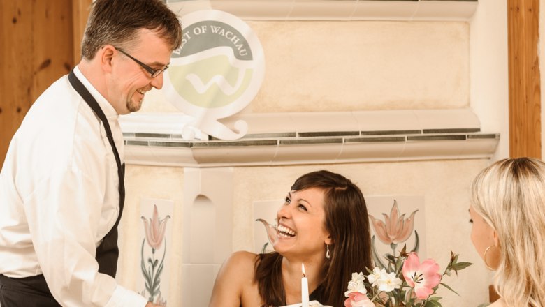Fürnbergstub'n Landhotel Wachau, © BEST WESTERN Landhotel Wachau A waiter serves two women in a cozy restaurant. One woman laughs as she accepts the food. The table is elegantly set.