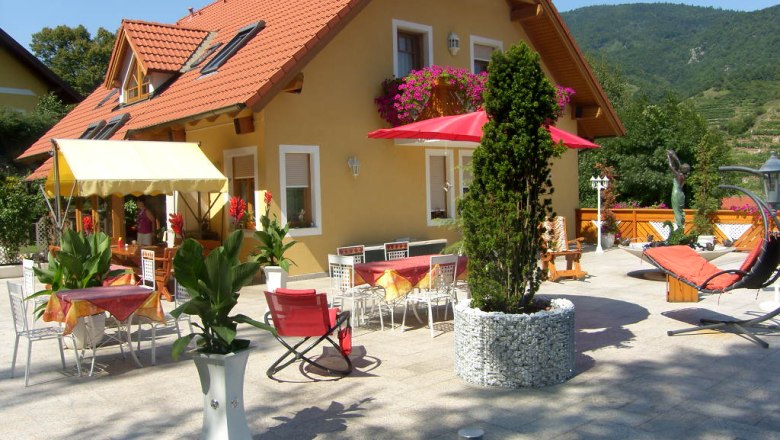 Gästehaus "Zur schönen Aussicht", © Elisabeth Wegscheider Gästehaus mit Terrasse, Tischen, Stühlen und Sonnenschirmen, umgeben von Pflanzen und Bergen im Hintergrund.