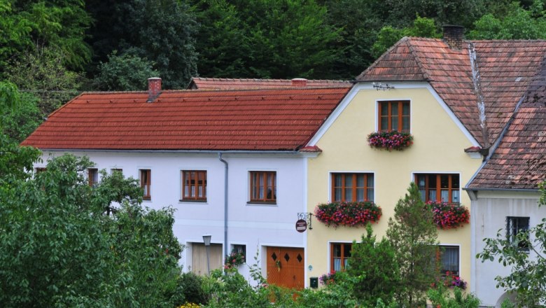 A traditional house with red roof tiles and flower boxes in front of the windows, surrounded by green vegetation.