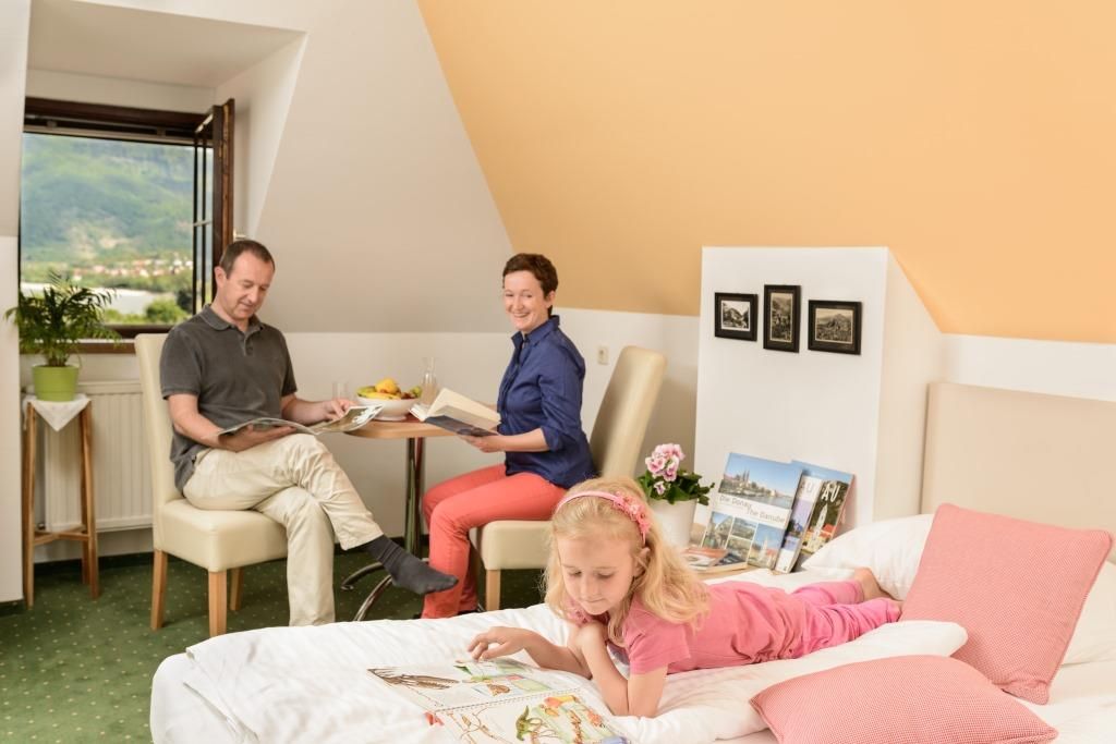Familie in einem gemütlichen Zimmer, Eltern lesen am Tisch, Kind liegt auf dem Bett mit einem Buch.