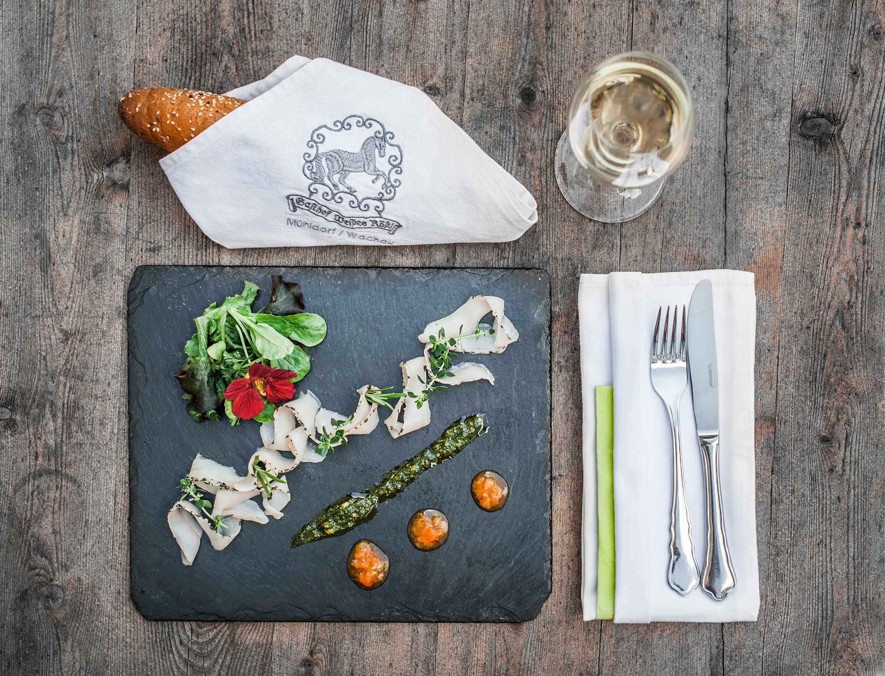 Elegante Vorspeise auf Schieferplatte mit Brot, Besteck und Weinglas auf Holztisch.