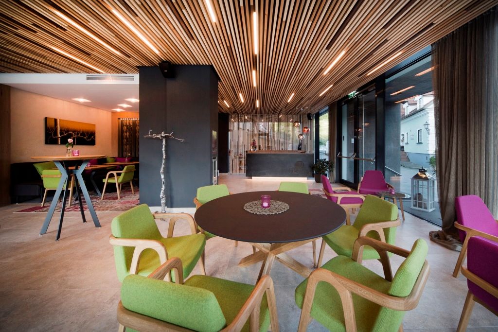 Innenansicht einer modernen Hotel-Lounge mit Holzelementen, grünen und lila Stühlen und einem runden Tisch.