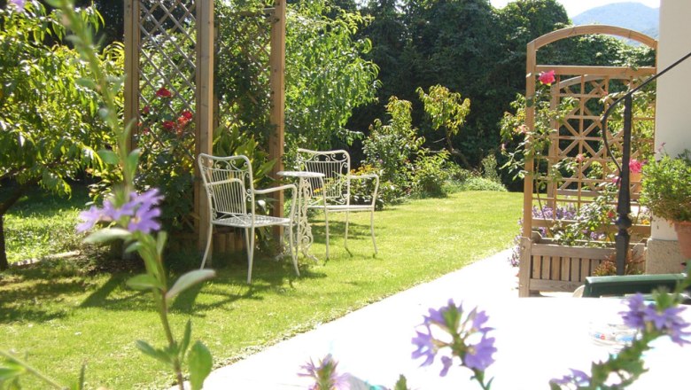 A sunny garden with white metal chairs, a table and flowering plants.