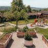 A playground with a slide and climbing frame next to raised beds in a garden with wooden huts in the background.