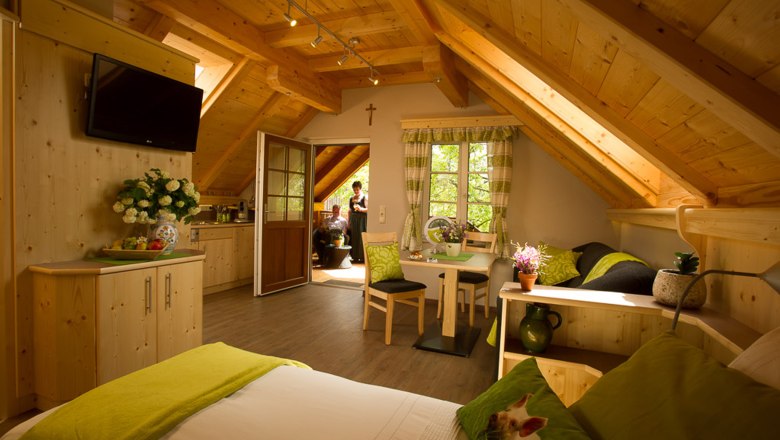 Cozy vacation apartment with wooden furnishings, sloping ceilings and green accents. Two people can be seen on a balcony in the background.