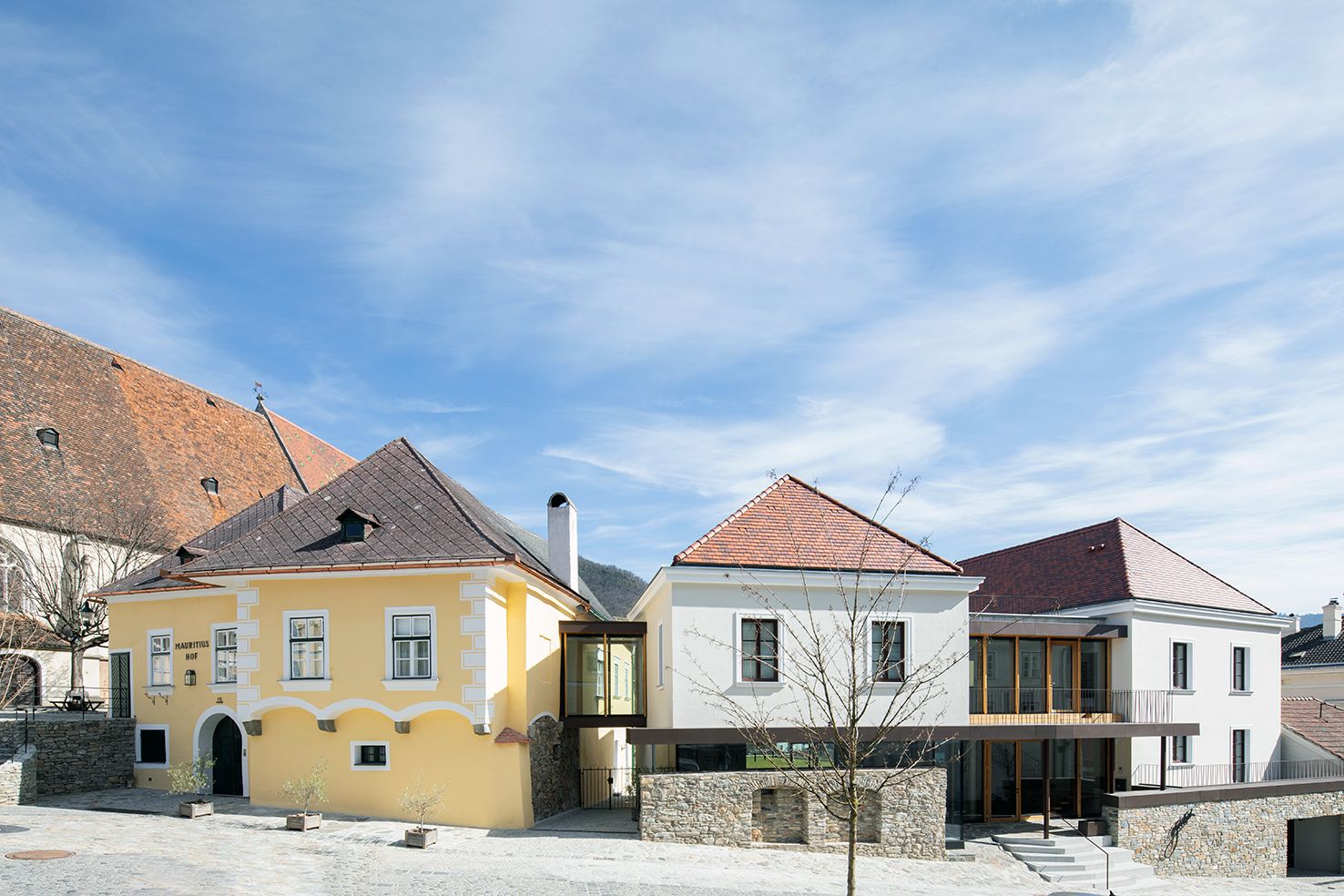 Zwei Gebäude mit gelber und weißer Fassade, verbunden durch einen modernen Glasgang, unter blauem Himmel.