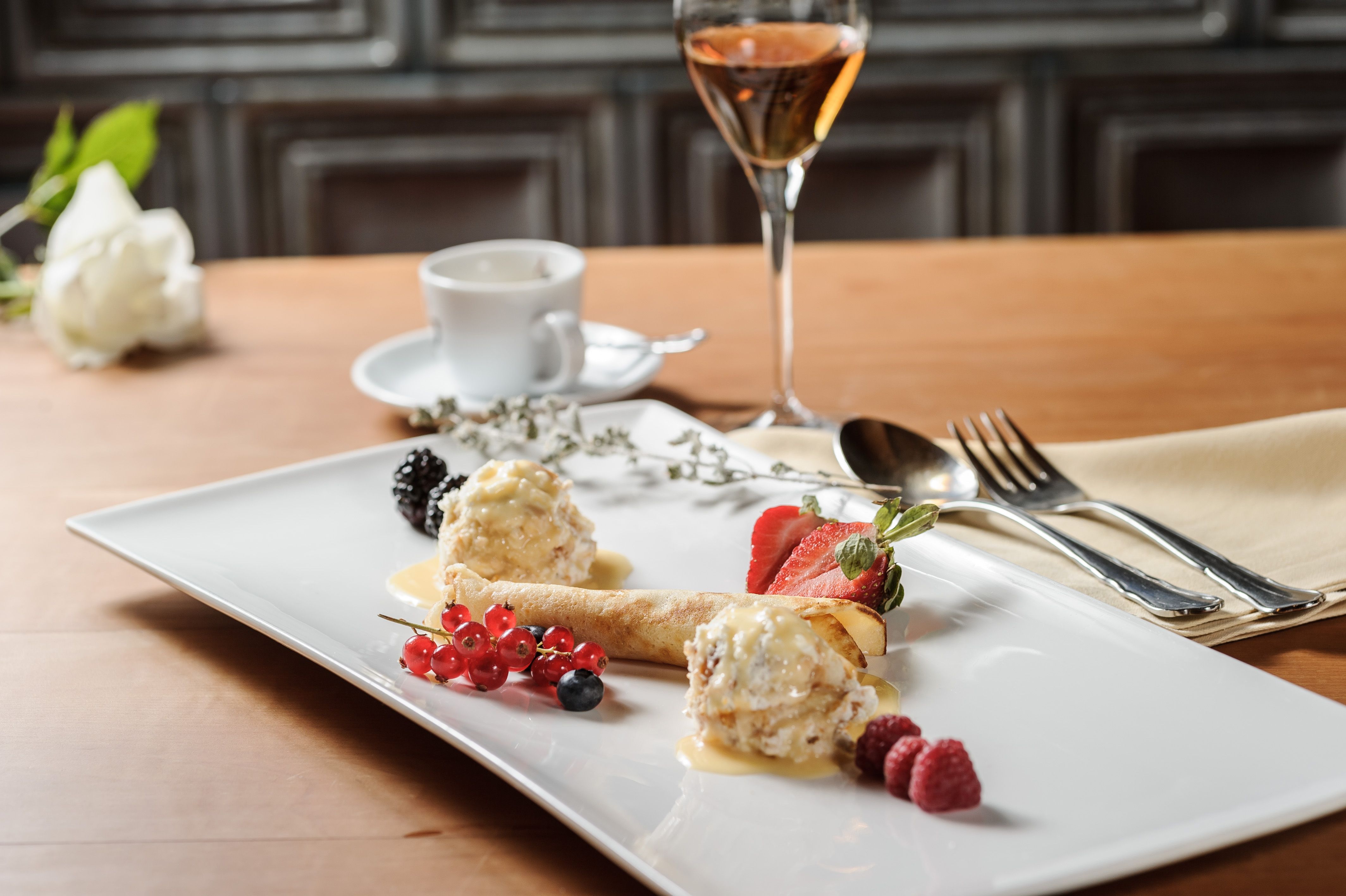 Elegantes Dessert mit Früchten und Eis auf einem weißen Teller, daneben ein Glas Wein und eine Tasse Kaffee.