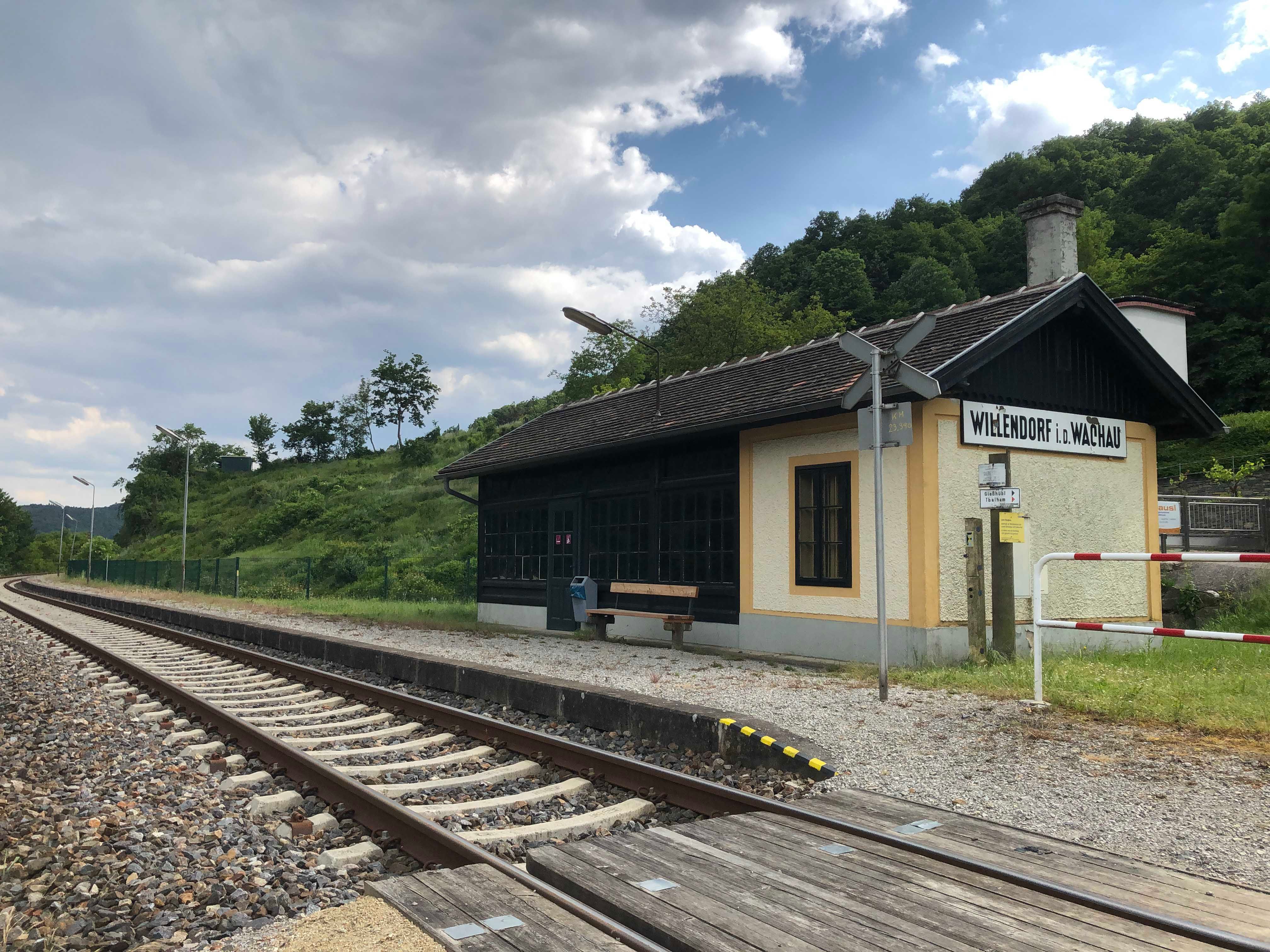 Kleiner Bahnhof in ländlicher Umgebung mit Gleisen und einem Wartehäuschen.