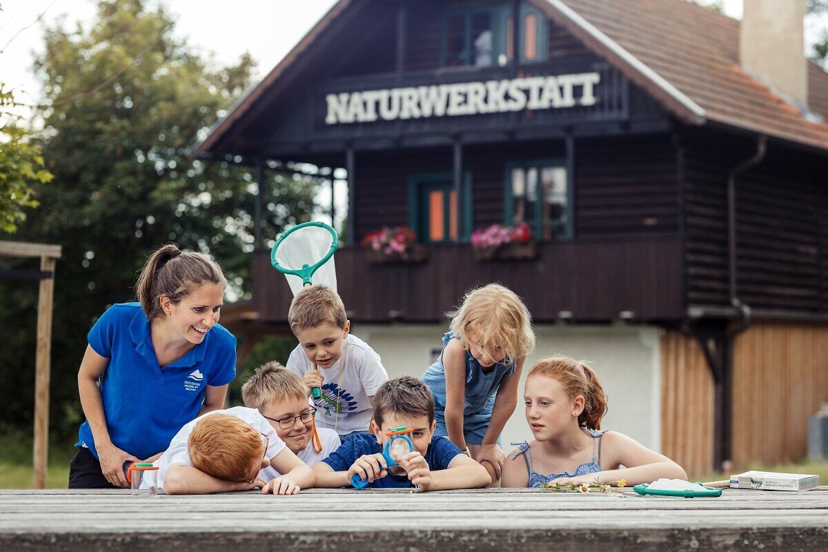 Eine Gruppe fröhlicher Kinder erkundet die Natur und entdeckt die Wunder der Umgebung. Unter der Anleitung einer engagierten Betreuerin lernen sie spielerisch über die Tier- und Pflanzenwelt. Die Atmosphäre ist geprägt von Neugier und Freude, während die Sonne sanft auf die Szene scheint.