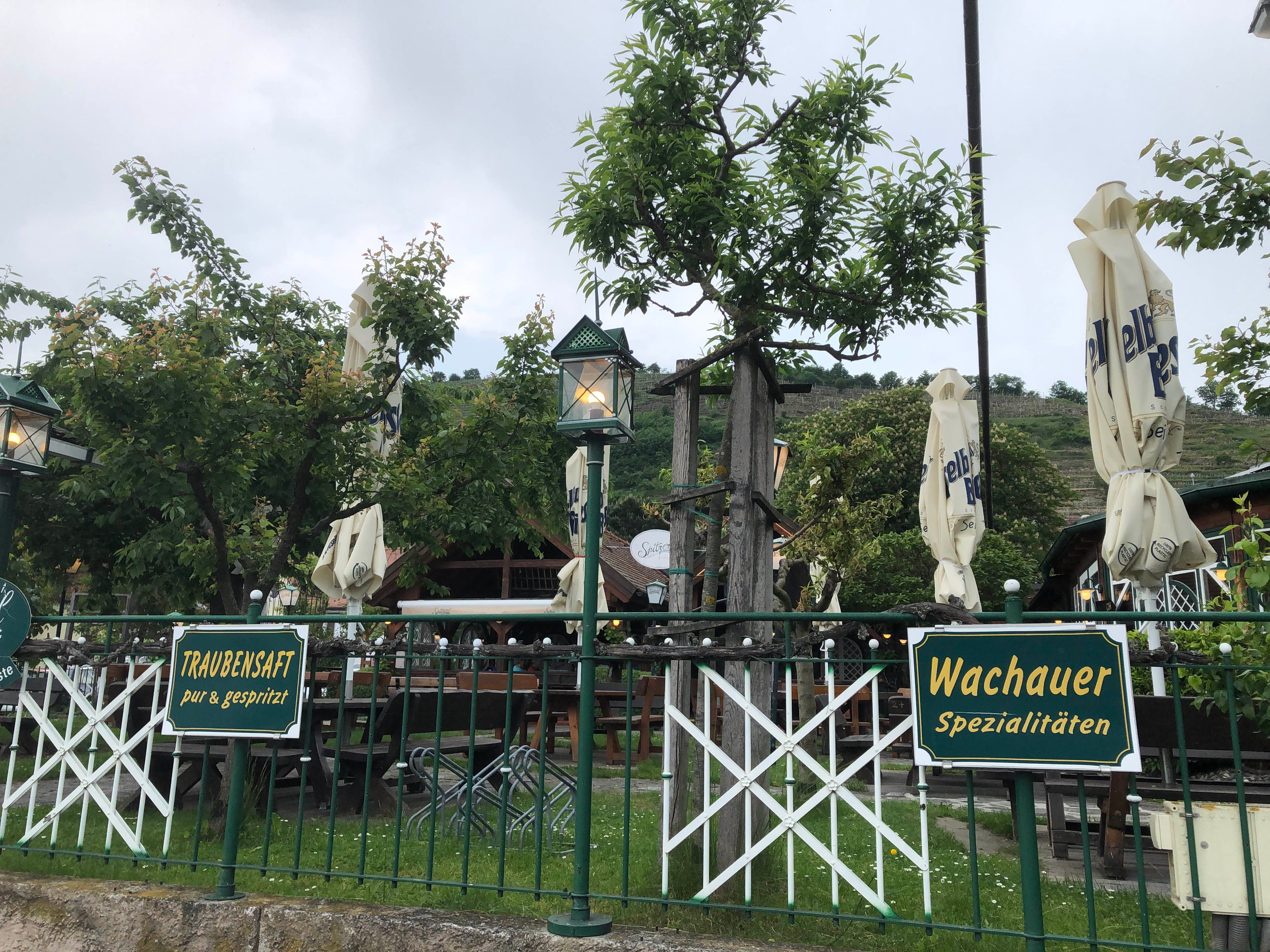 Gastgarten mit Schildern für Traubensaft und Wachauer Spezialitäten, umgeben von Bäumen und geschlossenen Sonnenschirmen.