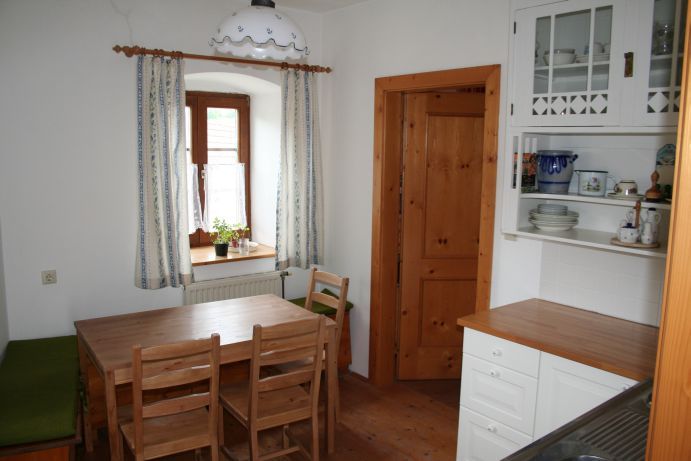 Kleine Küche mit Holzmöbeln, Esstisch, Stühlen und einem Fenster mit Vorhängen.