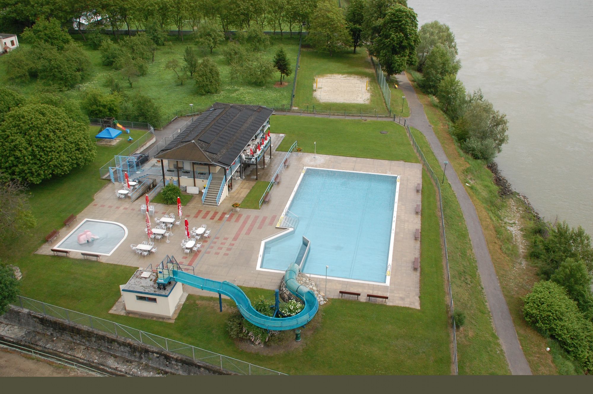 Luftaufnahme eines Freibads mit Schwimmbecken, Rutsche und Liegewiese am Flussufer.