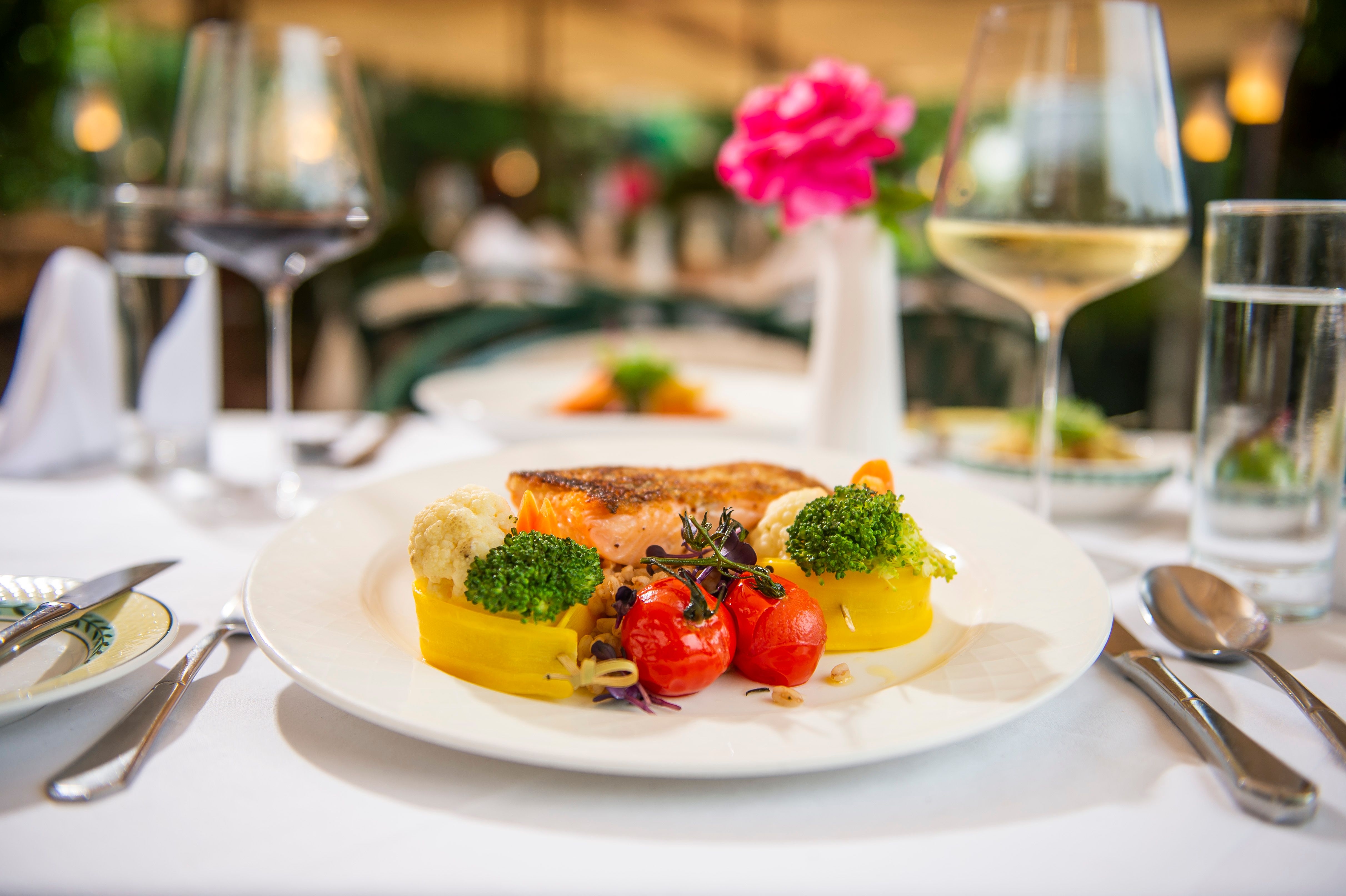 Ein Teller mit gegrilltem Lachs, Brokkoli, Blumenkohl, gelber Paprika und Kirschtomaten, auf einem gedeckten Tisch mit Weingläsern.