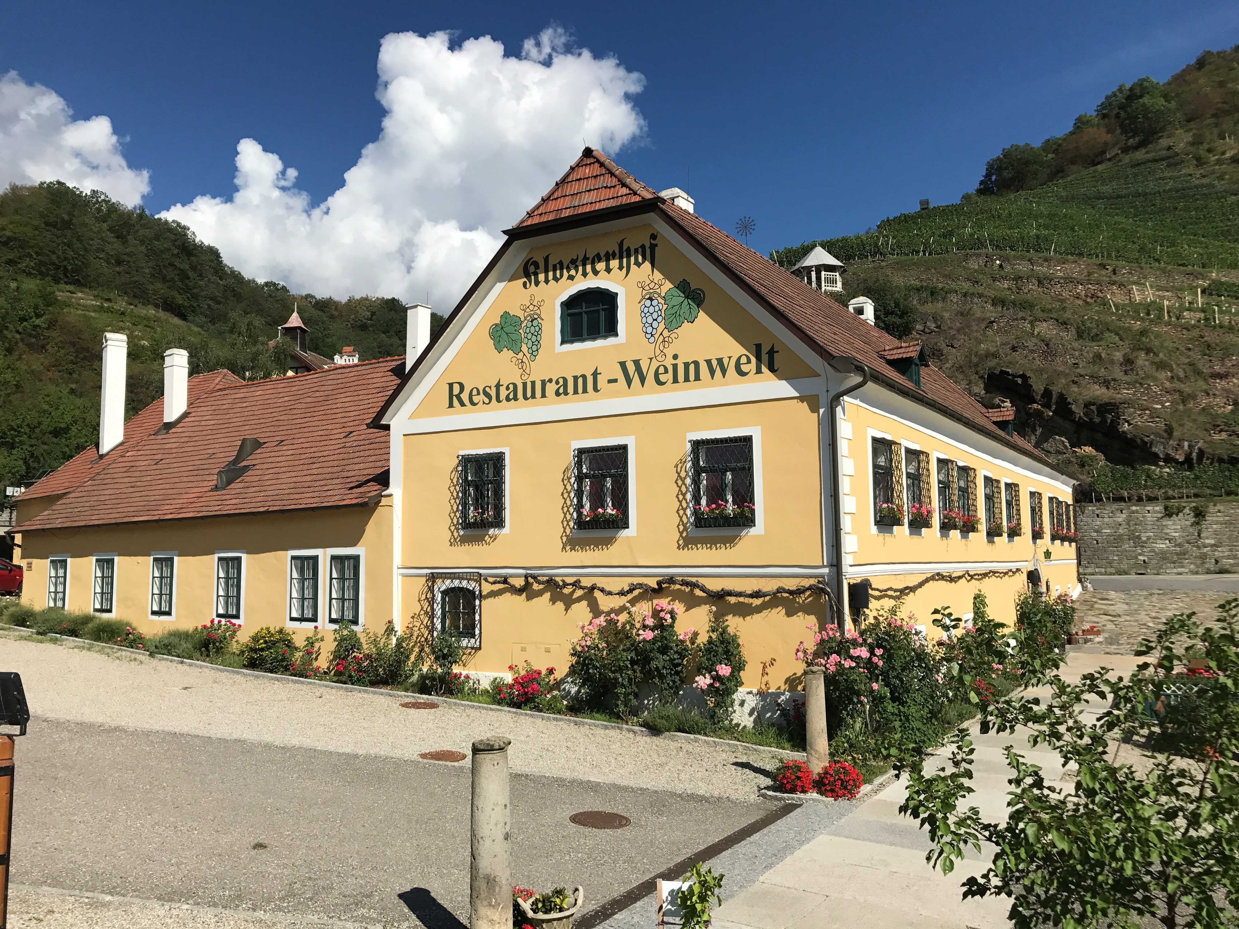 Außenansicht eines gelben Gebäudes mit der Aufschrift 'Klosterhof Restaurant-Weinwelt', umgeben von Blumen und Weinbergen.