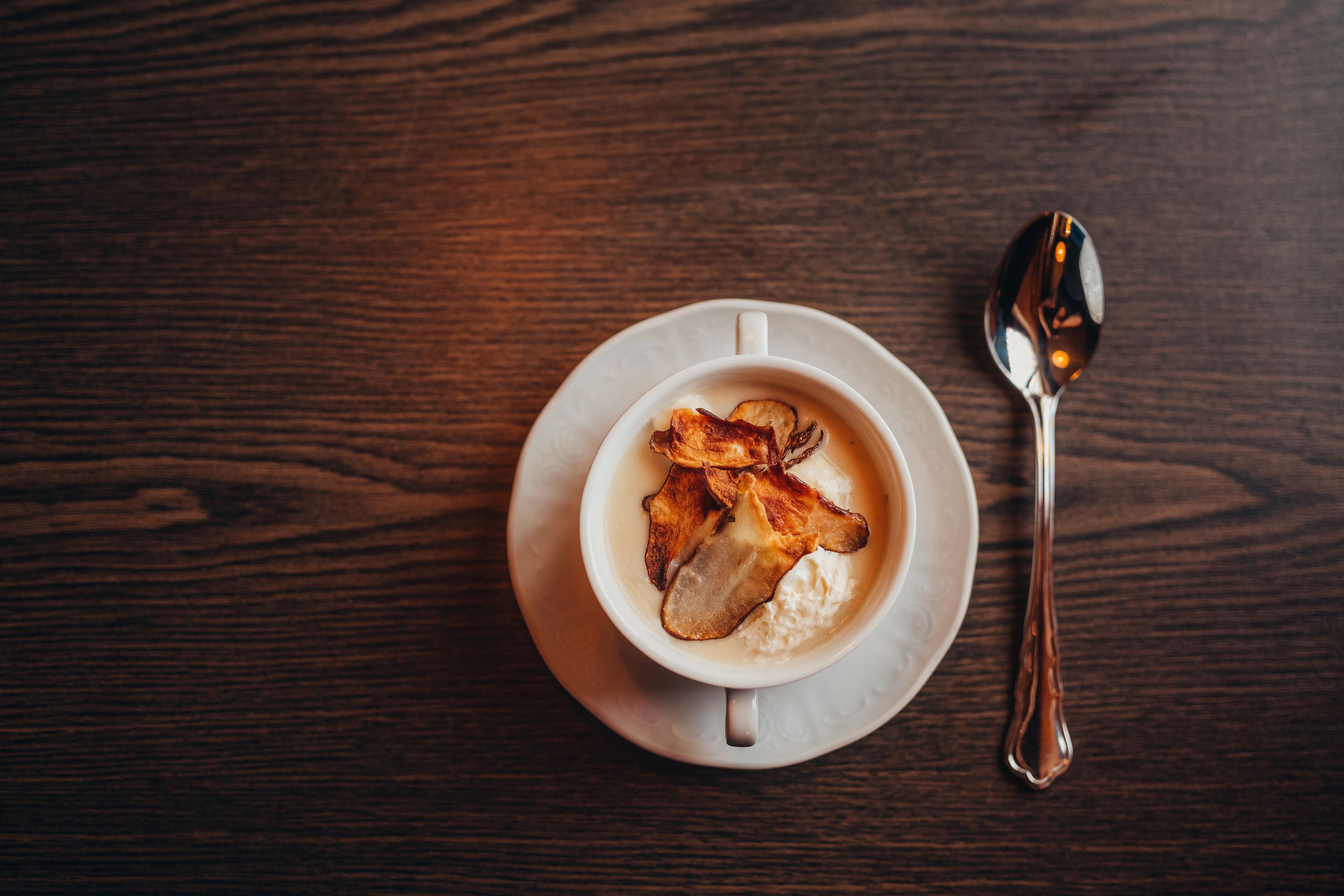 Eine Tasse Topinambur-Suppe mit Chips auf einem Holztisch, daneben ein Löffel.