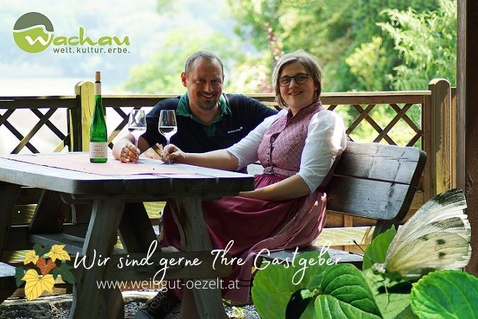 Ein Paar sitzt an einem Holztisch im Freien mit Wein und Gläsern. Im Hintergrund ist Natur zu sehen. Text: "Wir sind gerne Ihre Gastgeber".