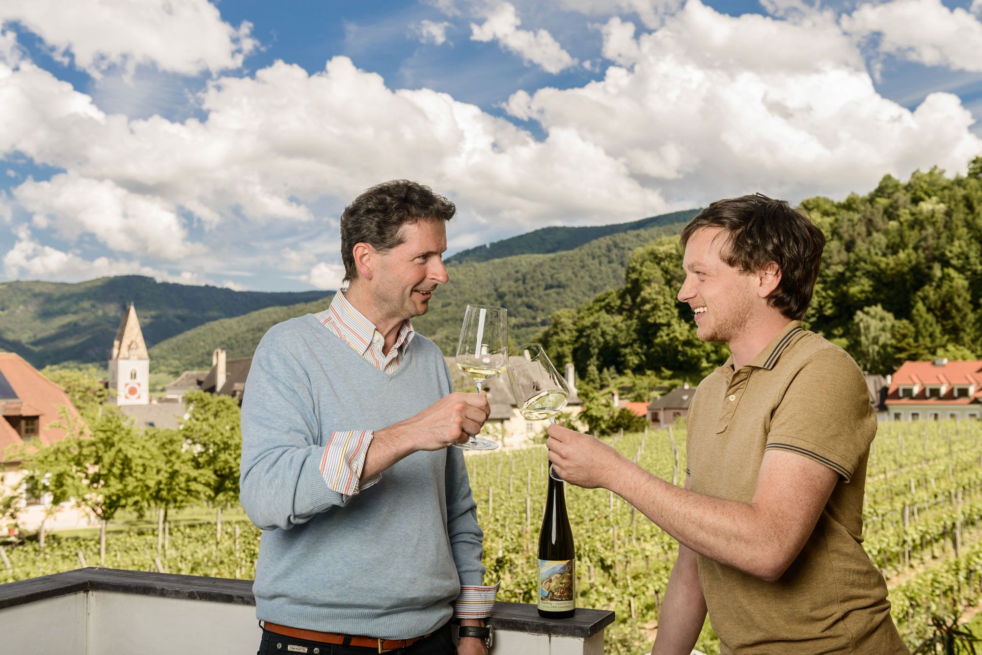 Zwei Männer stoßen mit Weingläsern an, im Hintergrund Weinberge und ein Kirchturm.