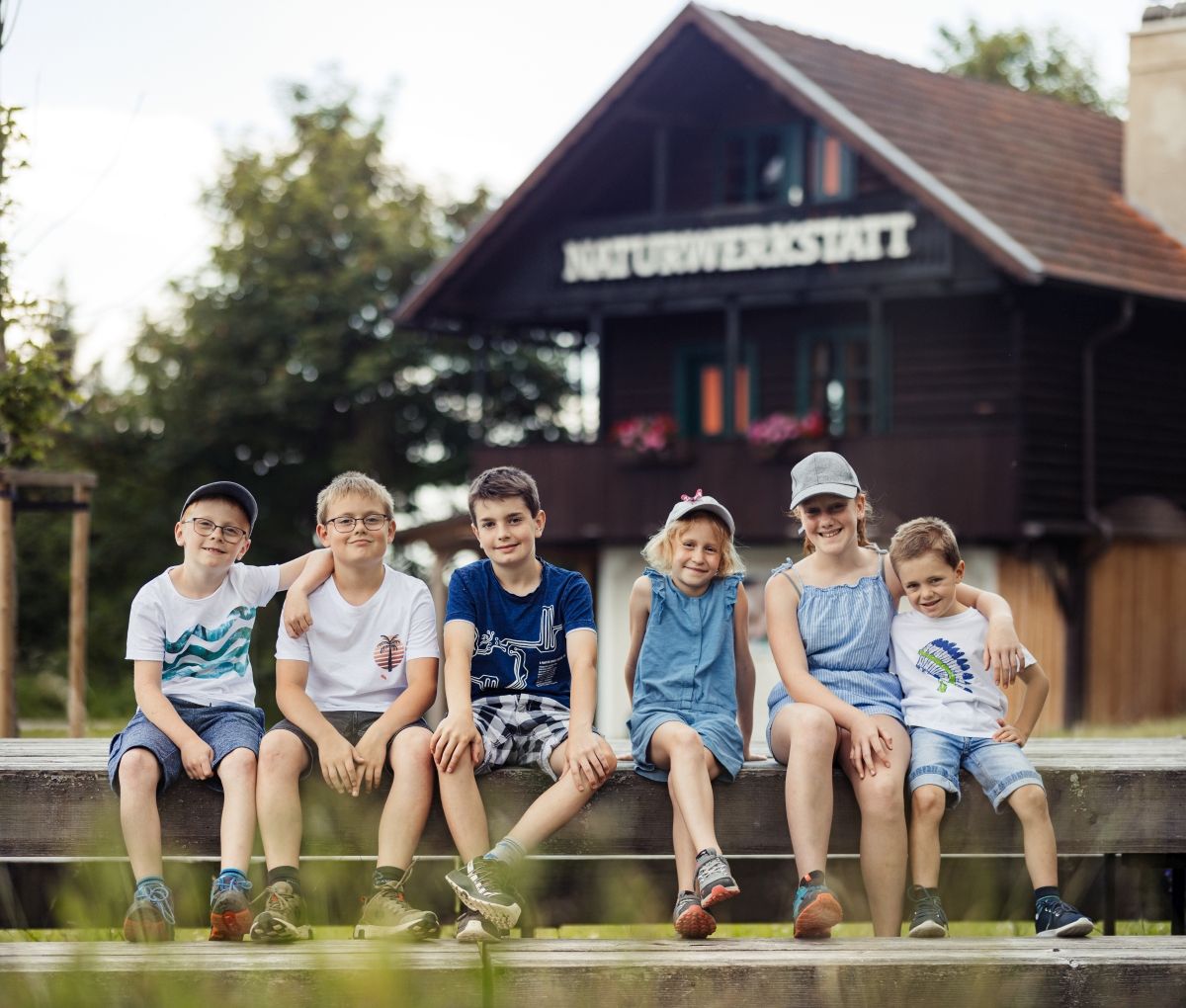 Eine Gruppe fröhlicher Kinder erkundet die Natur und entdeckt die Wunder der Umgebung. Unter der Anleitung einer engagierten Betreuerin lernen sie spielerisch über die Tier- und Pflanzenwelt. Die Atmosphäre ist geprägt von Neugier und Freude, während die Sonne sanft auf die Szene scheint.