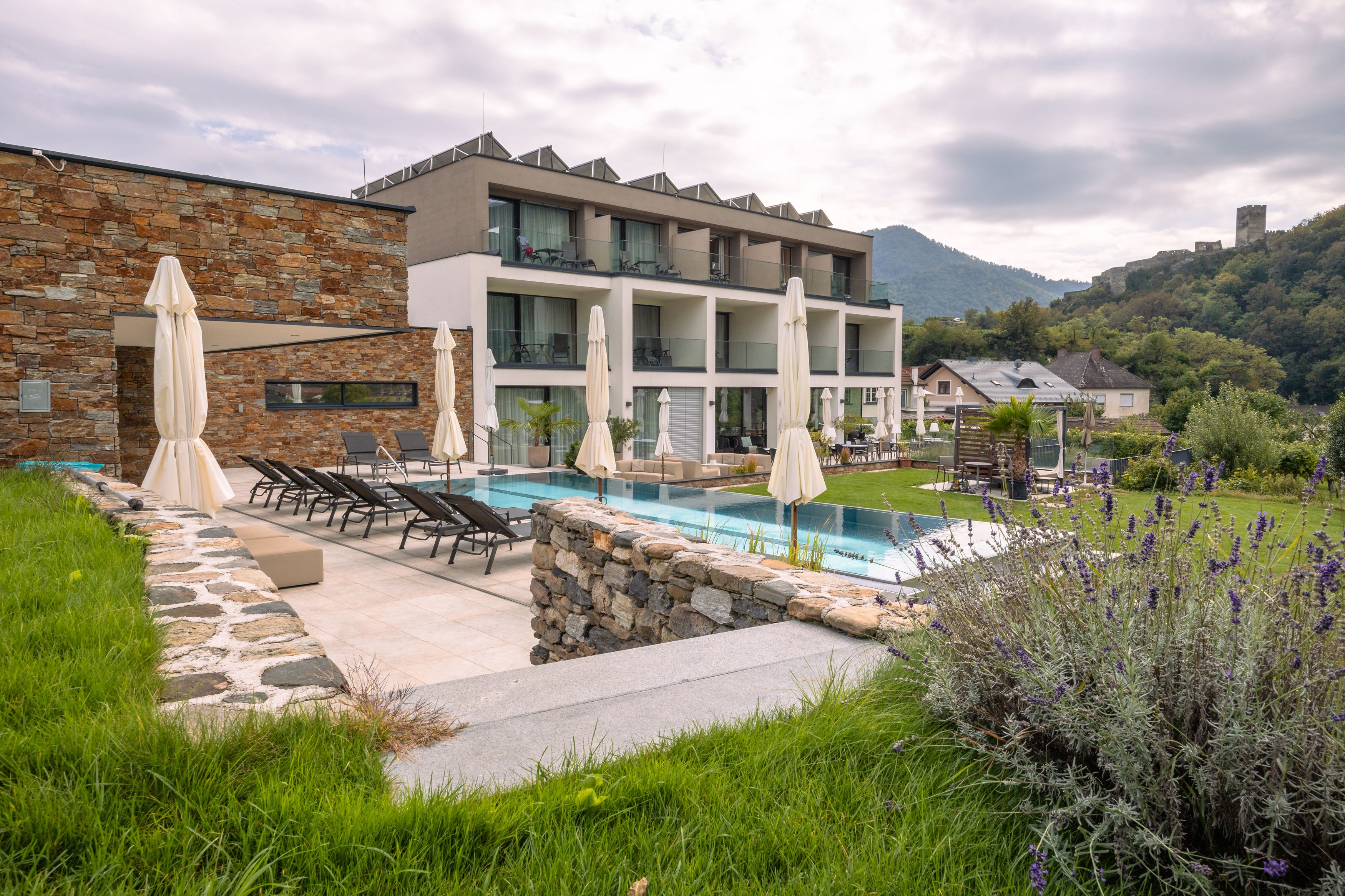 Außenansicht eines modernen Boutiquehotels mit Pool und Liegestühlen, umgeben von grüner Landschaft und Hügeln.