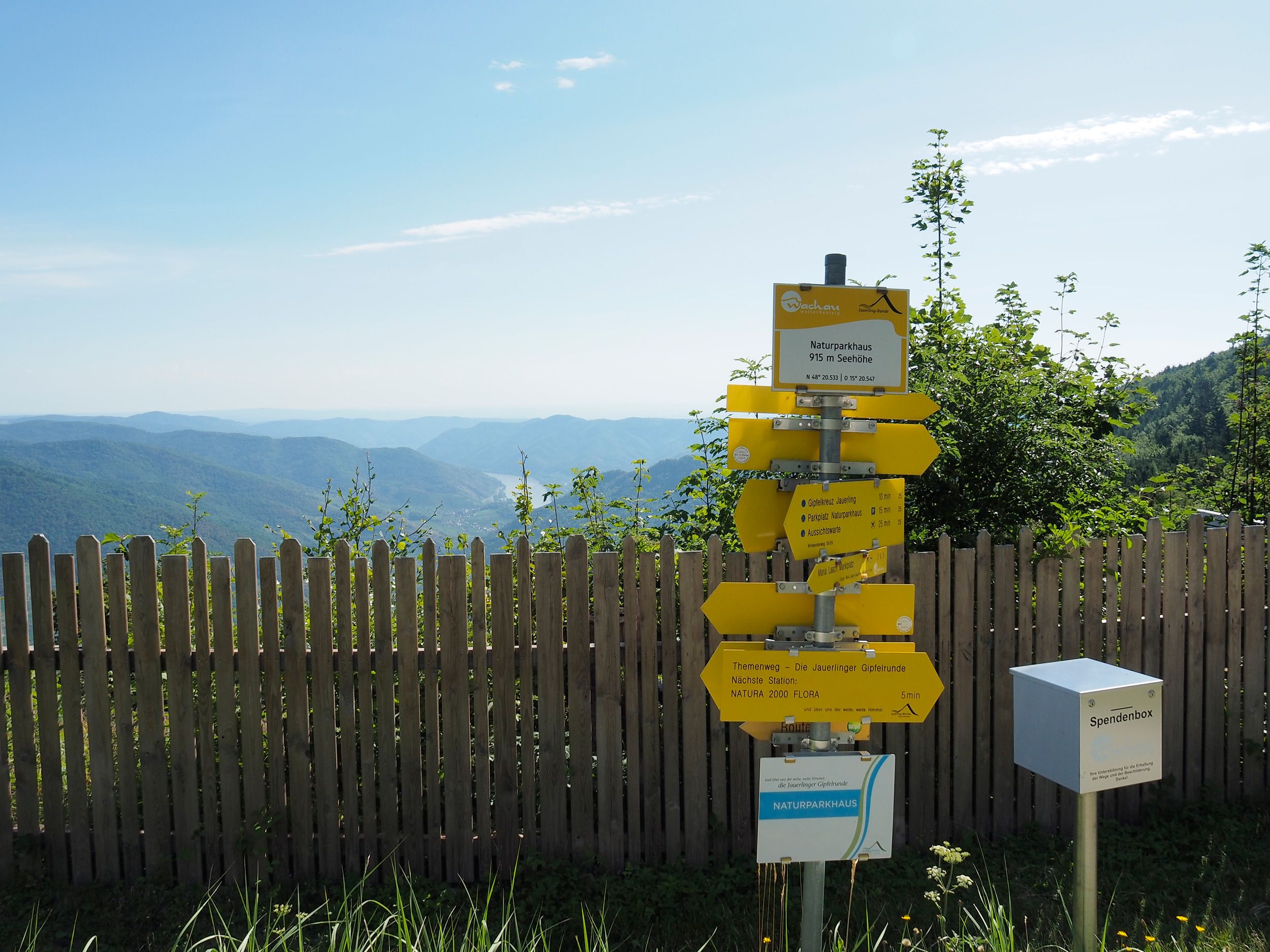 Wegweiser am Jauerling mit Blick auf bewaldete Hügel und blauen Himmel.