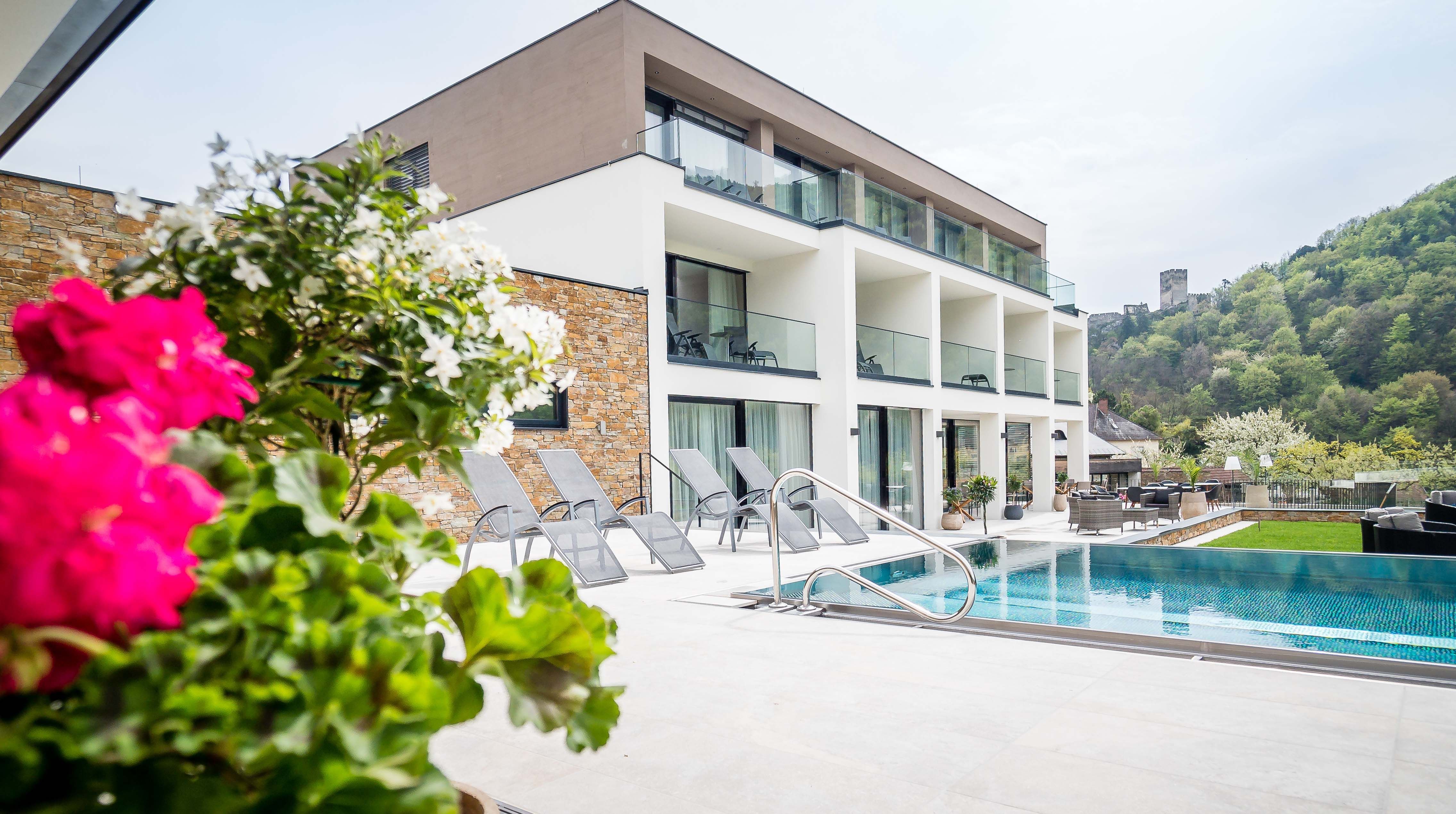 Modernes Gebäude mit Pool und Liegestühlen, umgeben von grüner Landschaft und Burg im Hintergrund.