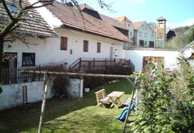 Ein ländlicher Hof mit weißen Gebäuden, Holztisch und Stühlen im Garten, Schaukel und Rutsche im Vordergrund.