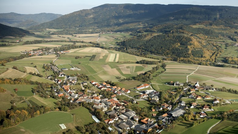 Luftaufnahme einer ländlichen Gemeinde mit Feldern und Hügeln im Hintergrund.