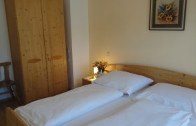 Ein gemütliches Schlafzimmer mit Doppelbett, Holzschrank, Nachttisch mit Blumen und einem Gemälde an der Wand.