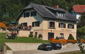 Ein zweistöckiges Haus mit der Aufschrift 'Haus Donaublick', umgeben von Blumen und Bäumen. Zwei Autos parken vor dem Haus.