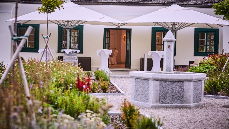 Ein steinerner Brunnen in einem Garten vor einem Geb&auml;ude mit wei&szlig;en Sonnenschirmen und gr&uuml;nen Fensterl&auml;den.