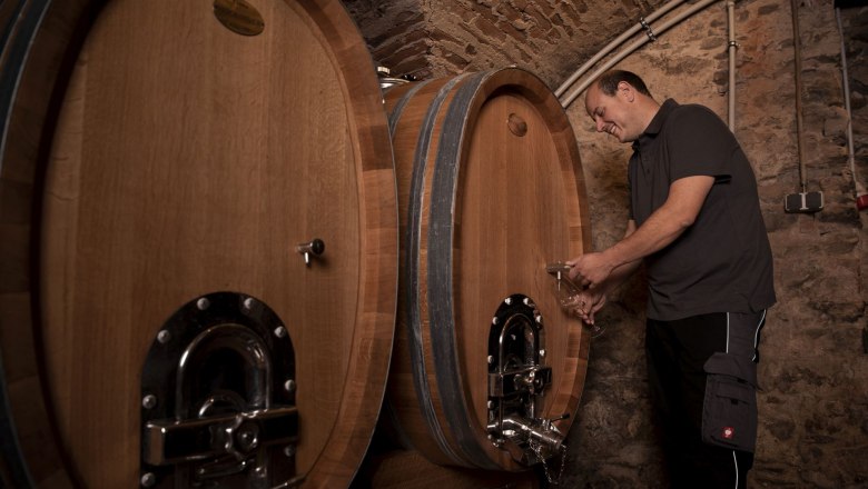 A man draws wine from a large wooden barrel in a wine cellar.