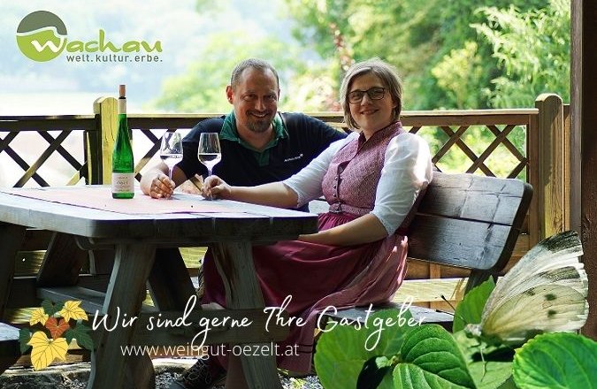 A couple sits at a wooden table outdoors with wine and glasses. Nature can be seen in the background. Text: "We are happy to be your hosts".