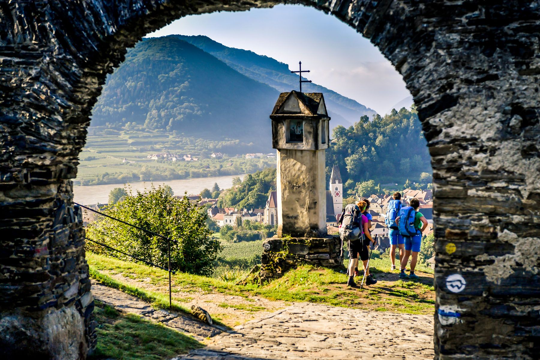 Blick durch ein steinernes Tor auf eine Landschaft mit Hügeln, einem Fluss und Wanderern.