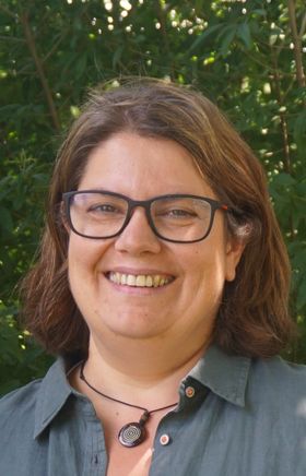 Eine Braunhaarige Brillenträgerin in Hemd lächelt. 