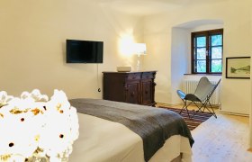 Cozy bedroom with bed, antique chest of drawers, armchair and wall-mounted TV.